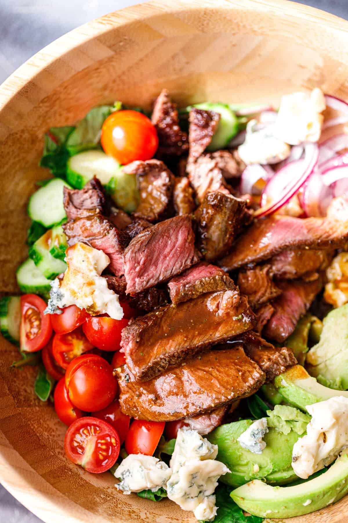 Close up of sliced steak, cherry tomatoes, avocado, and blue cheese over mixed greens.