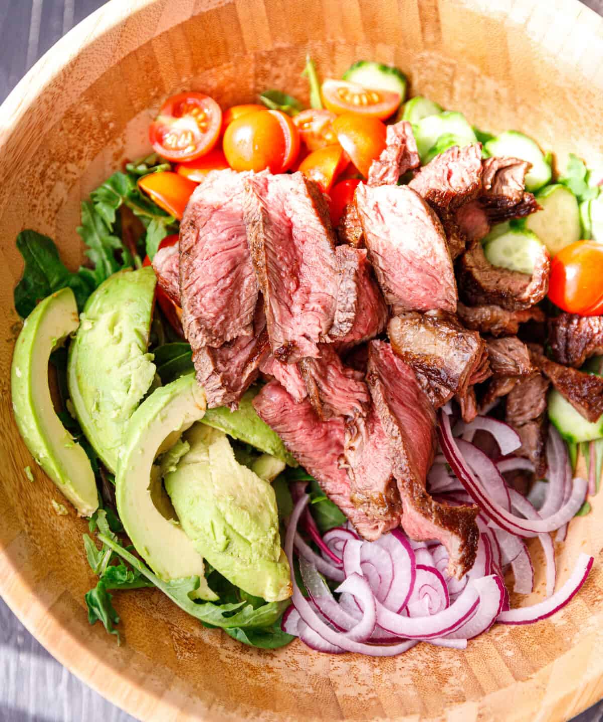Warm sliced steak layered over mixed greens, avocado, tomatoes, cucumbers, and red onion in a wooden bowl.