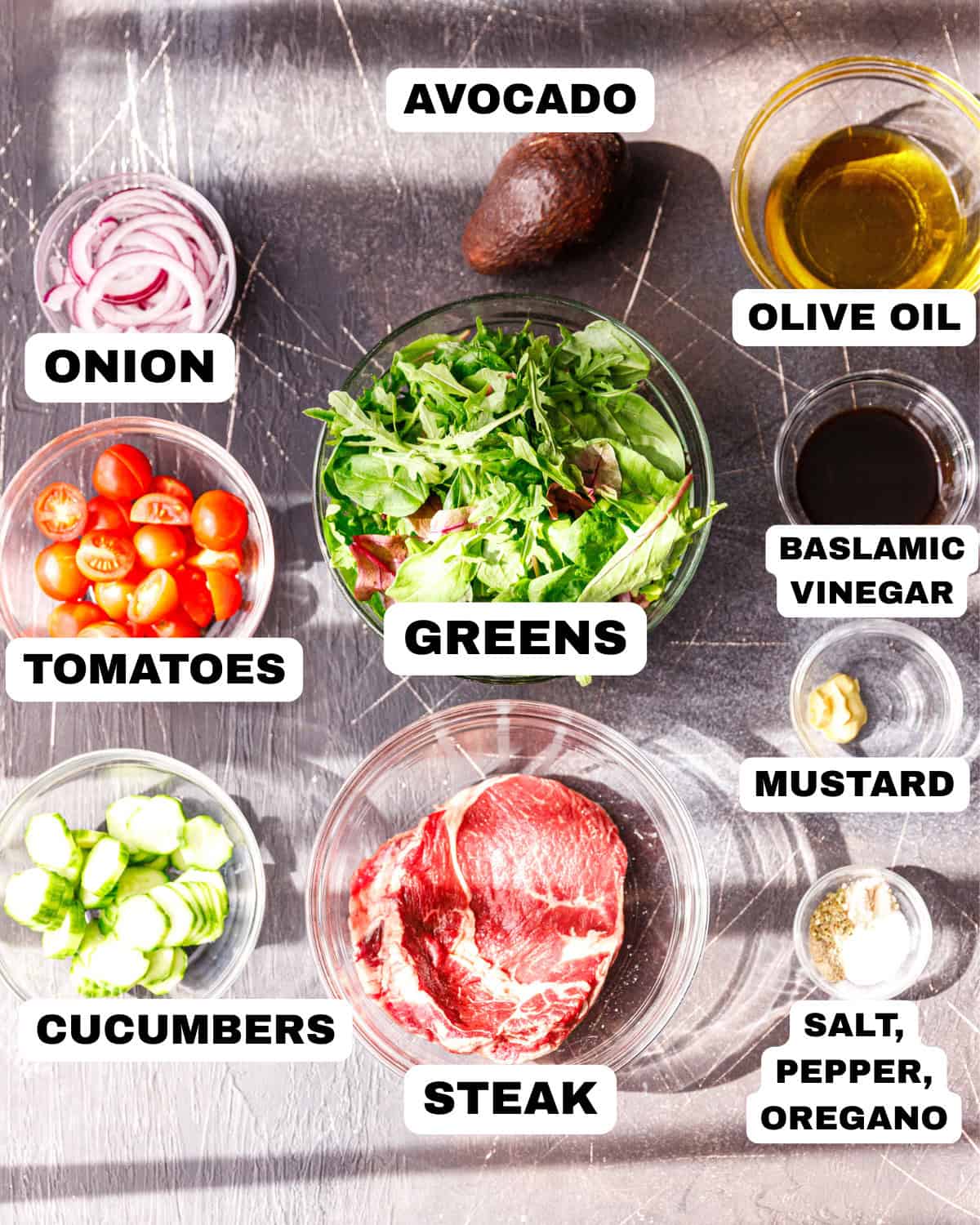 Steak salad ingredients laid out on a bright kitchen counter, including raw steak, mixed greens, cherry tomatoes, sliced cucumbers, thinly sliced red onion, an avocado, olive oil, balsamic vinegar, mustard, and simple seasonings.