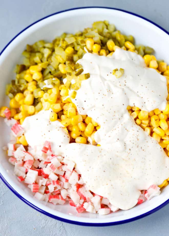 A white bowl with pickles, corn, imitation crab meat and yogurt dressing