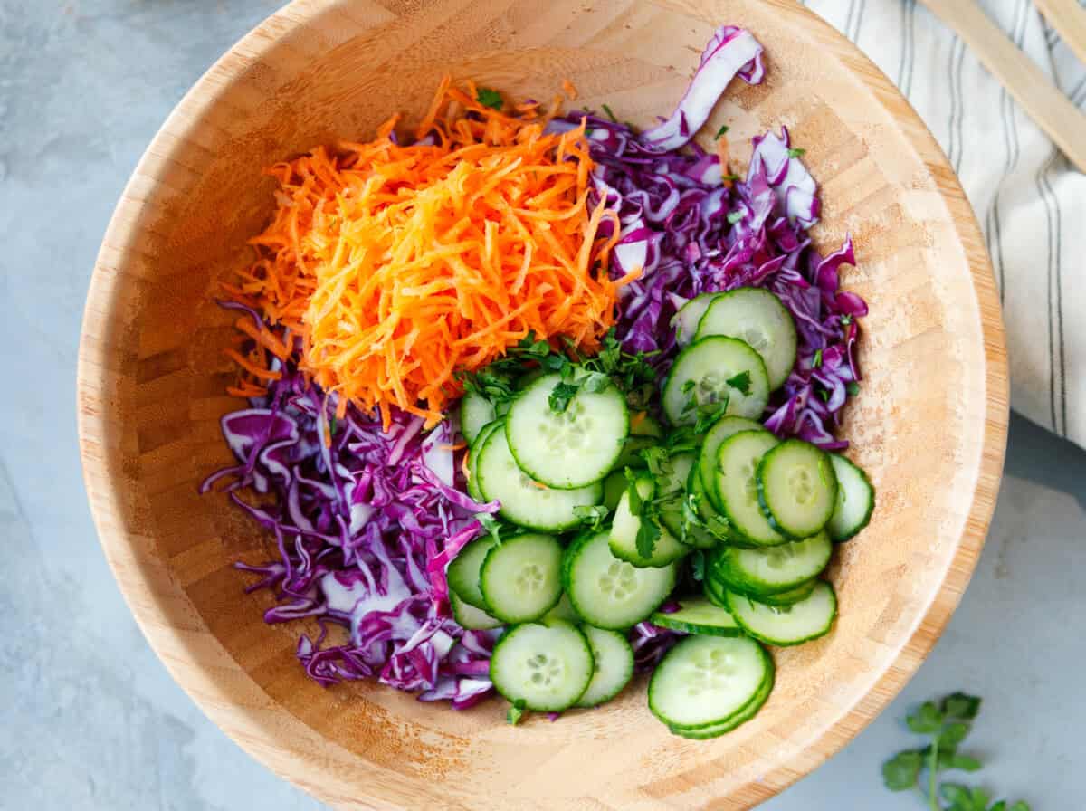 Shredded purple cabbage, grated carrots, sliced cucumbers and mixed seeds in wooden bowl before dressing