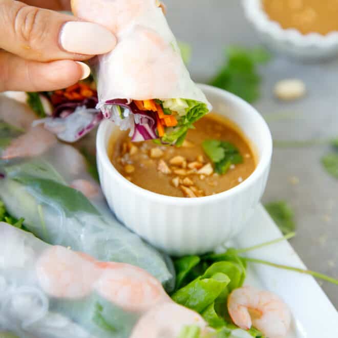 A hand holding shrimp spring roll over peanut dipping sauce
