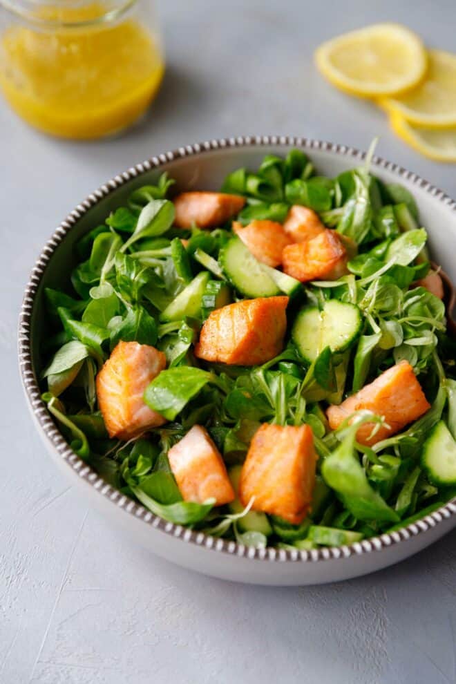 A bowl with greens, salmon and a jar with lemon dressing next to it.