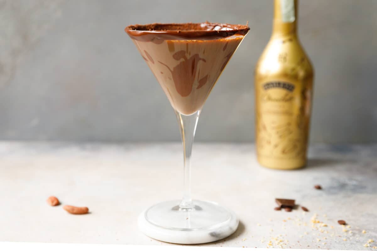A martini glass filled Baileys Chocolate Martini, on a marble coaster, with a bottle of Baileys Chocolat next to it.