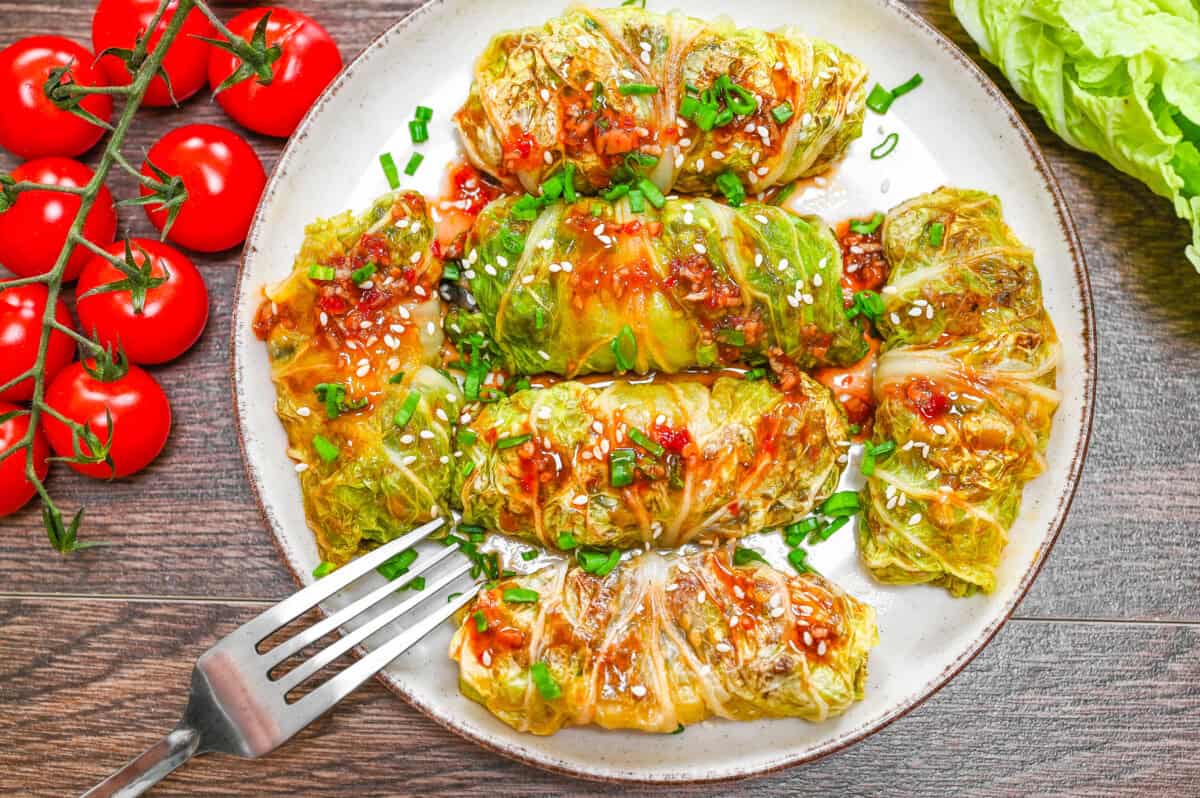 Plate of golden pan-seared cabbage rolls garnished with sesame seeds and scallions, served with a flavorful soy-based sauce and vine tomatoes on the side