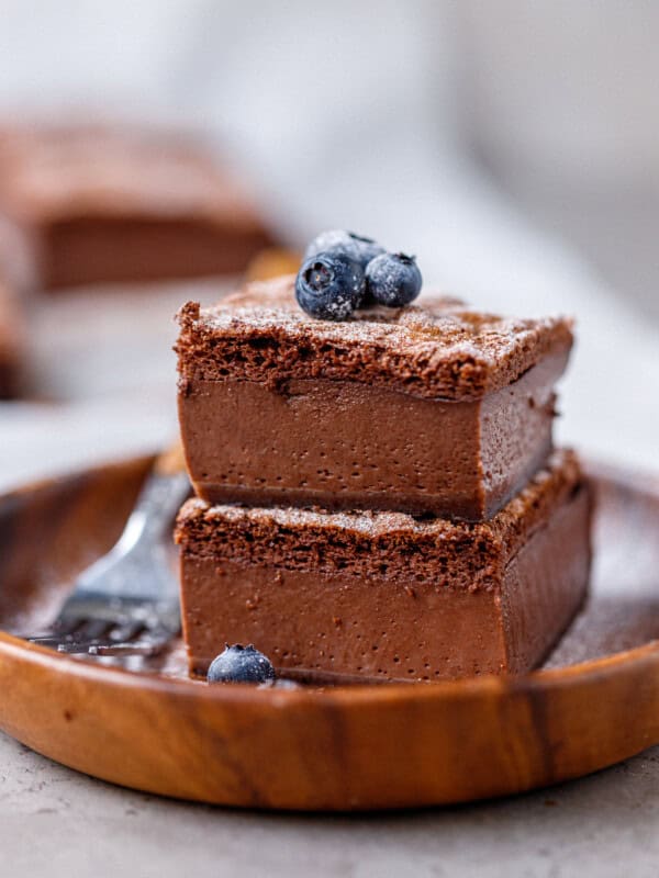 A close-up of a rich chocolate magic custard cake slice topped with fresh blueberries, showing its three distinct layers: fudgy base, creamy custard center, and airy top.