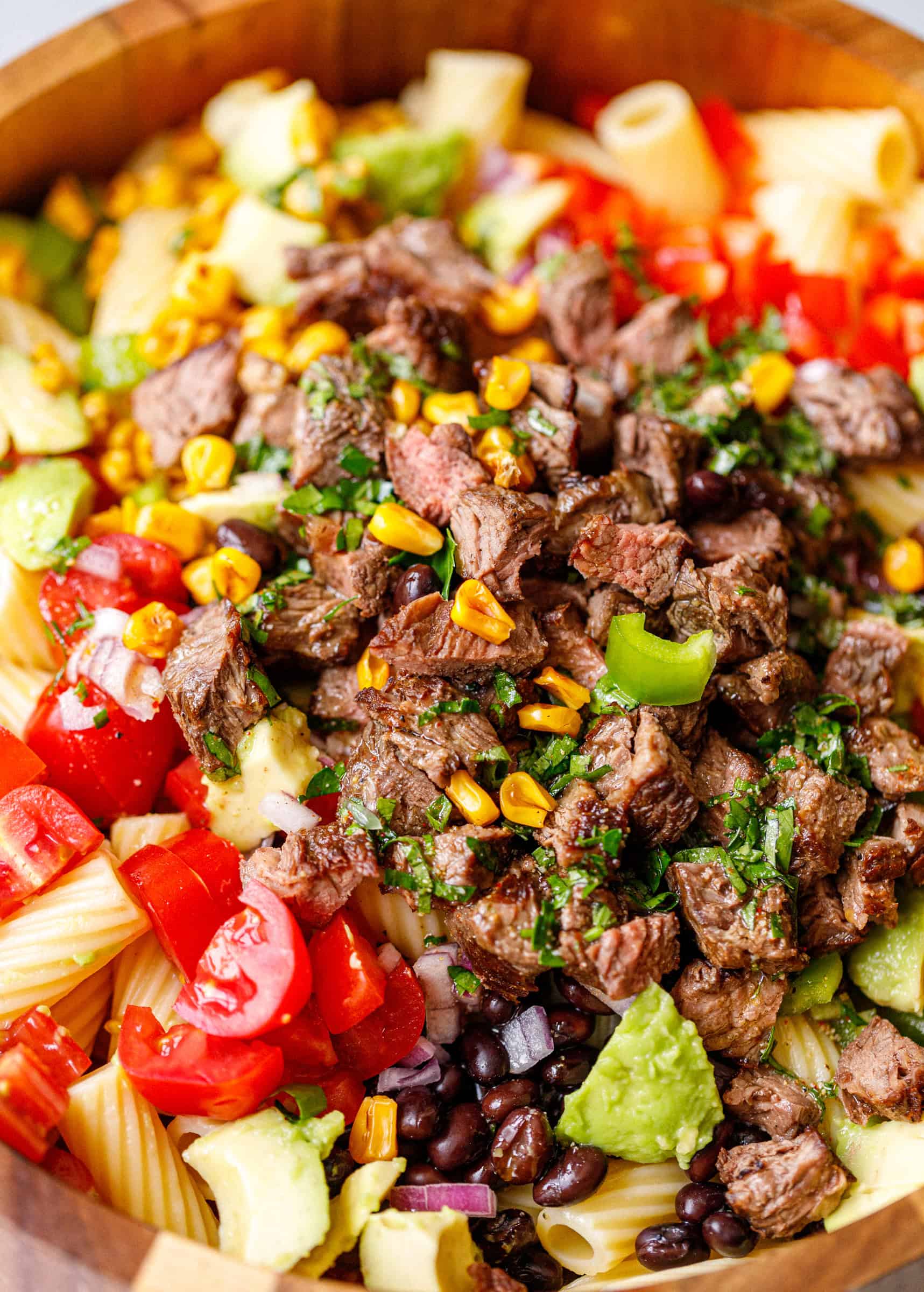 Close-up of steak pasta salad with black beans, avocado, tomatoes, corn, and rigatoni in a wooden bowl.