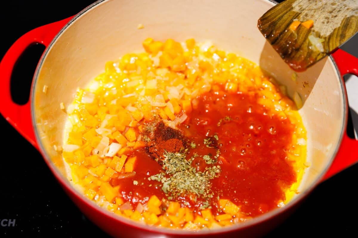 A Dutch oven on a stove top with onions, carrots, garlic, tomato sauce and seasoning being sautéed