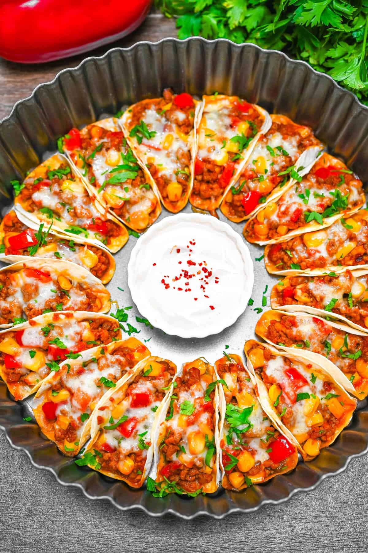 Overhead view of a full tray of mini tacos baked with cheese and vegetables, served with sour cream in the middle.