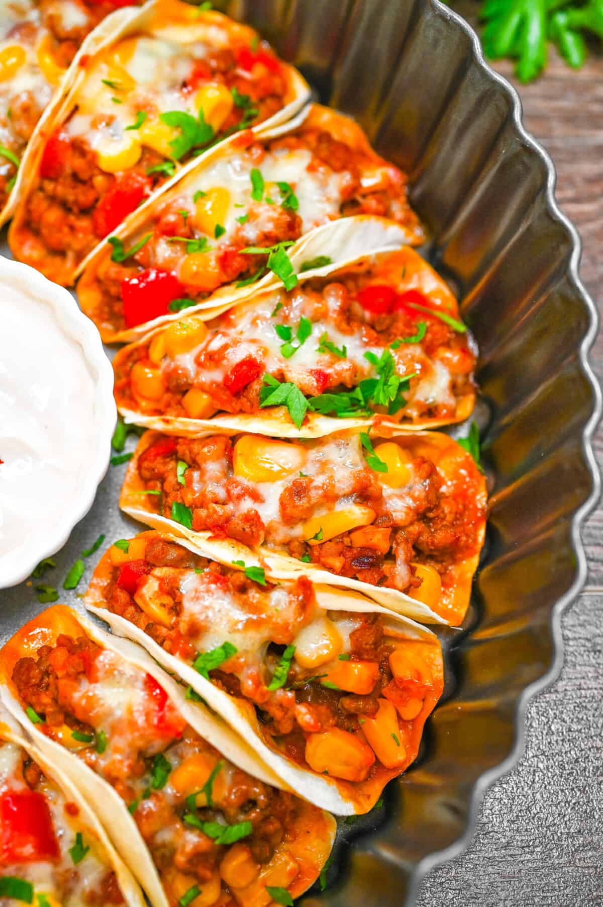 Mini beef tacos baked in a round tray with corn, peppers, and melted cheese, garnished with parsley, served alongside sour cream dip.
