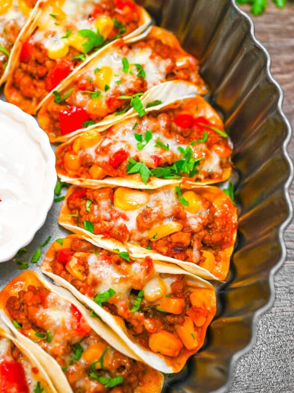 Mini beef tacos baked in a round tray with corn, peppers, and melted cheese, garnished with parsley, served alongside sour cream dip.