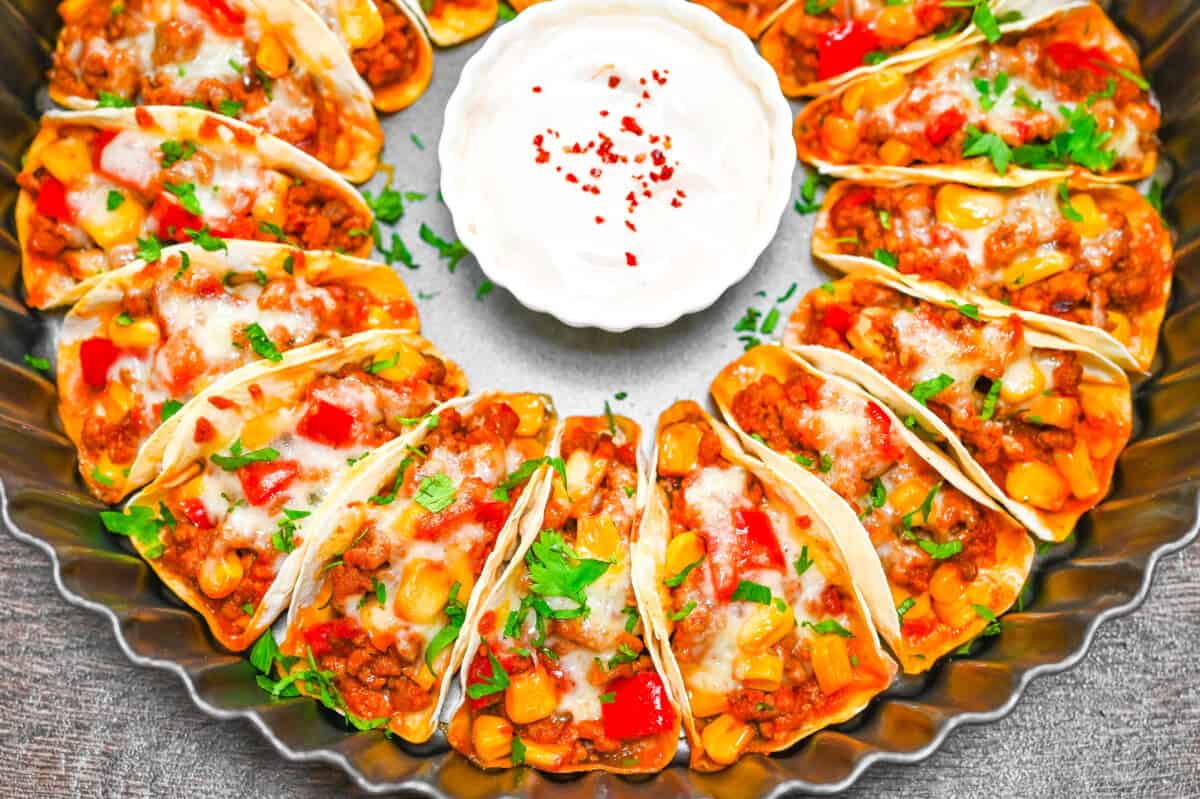 Round tray of mini tacos filled with beef, corn, and peppers, arranged in a circle with sour cream dip in the center.