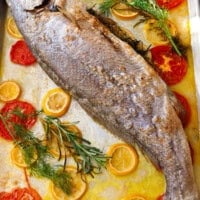 Whole roasted milokopi fish on a baking sheet with lemon slices, tomatoes, rosemary, and thyme.