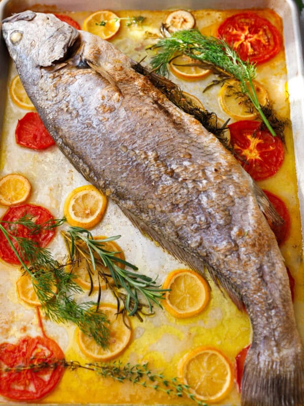 Whole roasted milokopi fish on a baking sheet with lemon slices, tomatoes, rosemary, and thyme.