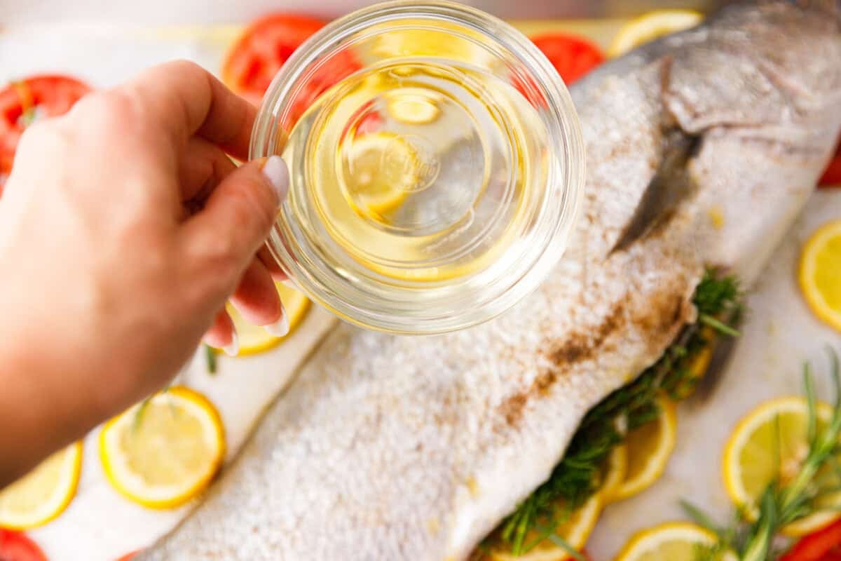 Hand holding a small bowl of white wine over a whole milokopi fish prepared with herbs and lemon slices.