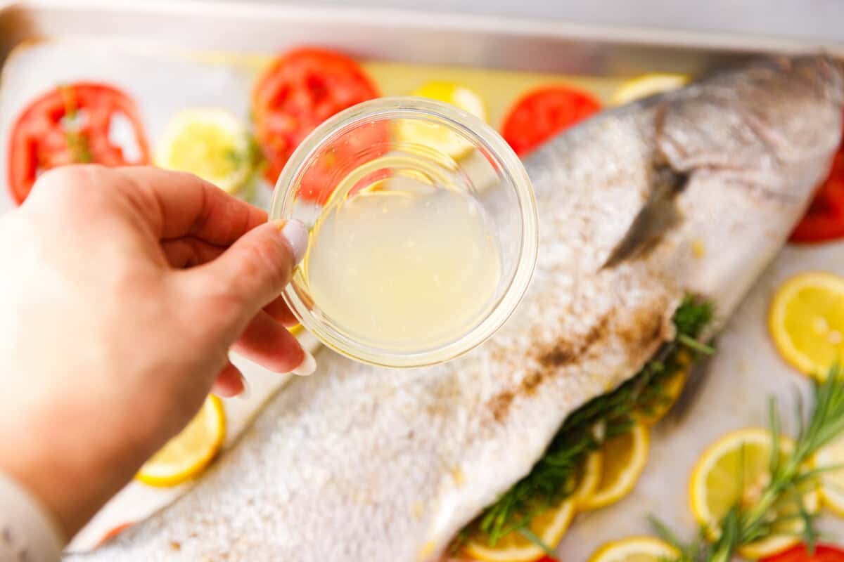 Hand holding a bowl of fresh lemon juice over a whole milokopi fish on a baking sheet with tomatoes, lemons, rosemary, and thyme.