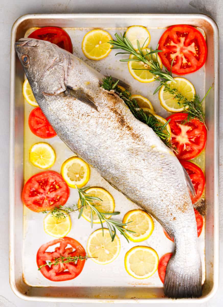 Whole seasoned milokopi fish placed on a baking sheet with lemon slices, tomato slices, rosemary, and thyme before roasting.