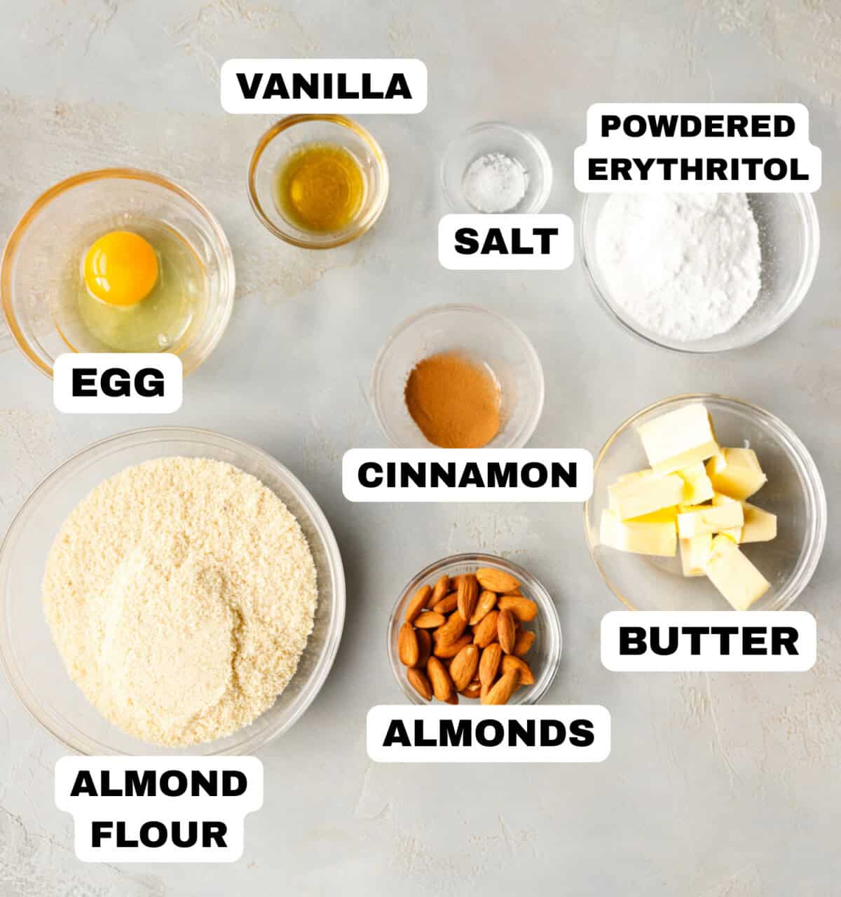 Keto almond cookie ingredients laid out on a light surface, including almond flour, butter, powdered erythritol, egg, vanilla, cinnamon, almonds, and salt.