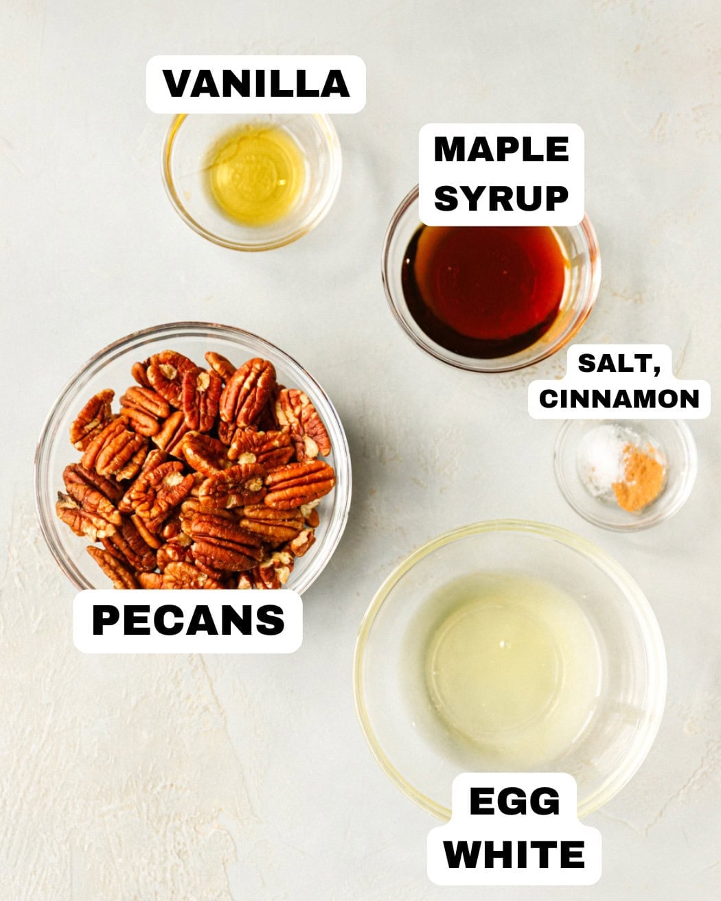 Ingredients for maple pecan cookies arranged in bowls, including pecans, egg white, maple syrup, vanilla, salt, and cinnamon