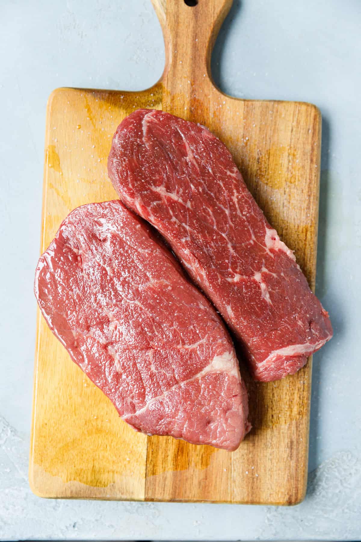 2 raw Denver steaks on a wooden board being salted before cooking