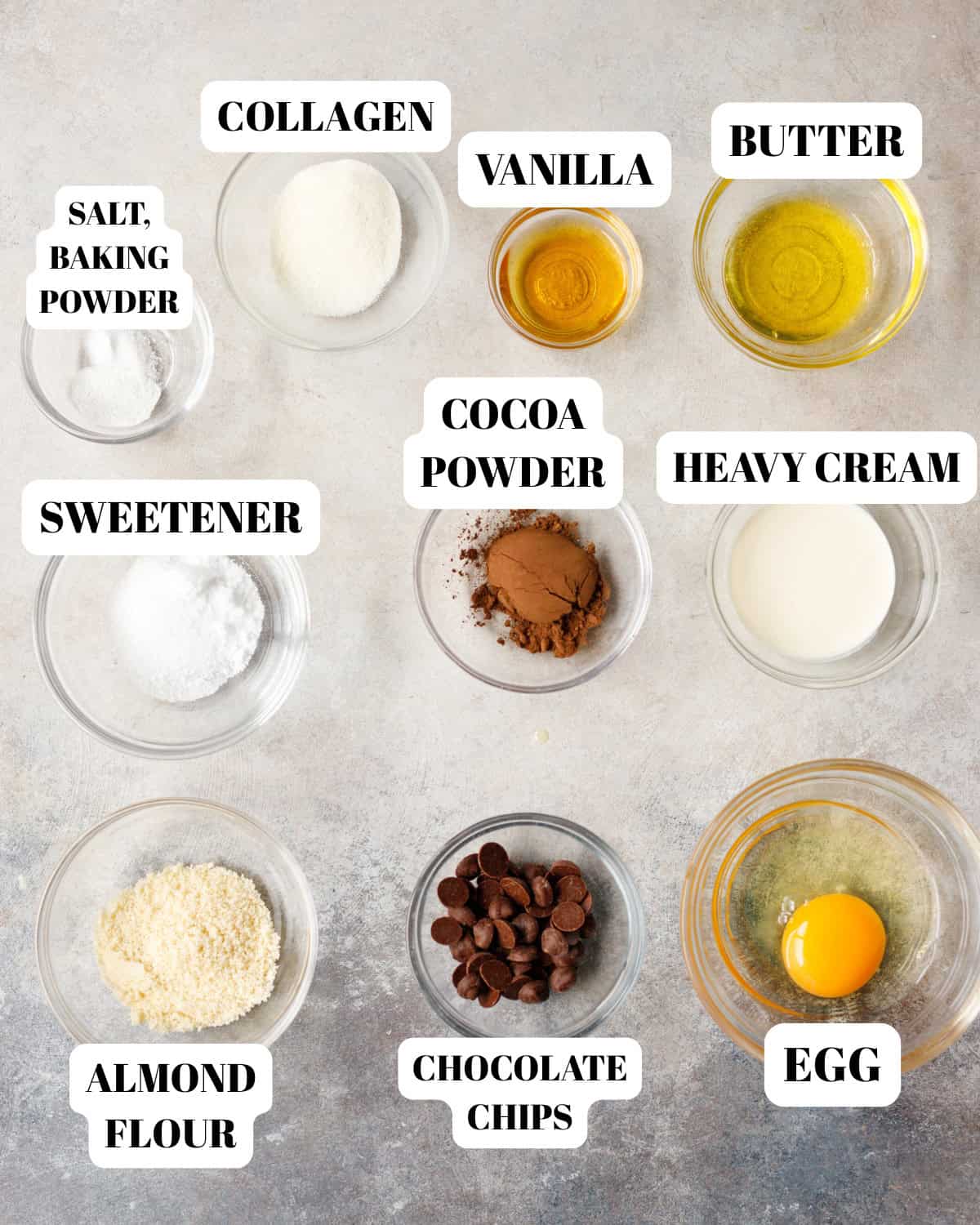 Chocolate collagen single serve cake ingredients including egg, cocoa powder, almond flour, sweetener, butter, heavy cream, vanilla, chocolate chips, baking powder and salt arranged in small bowls