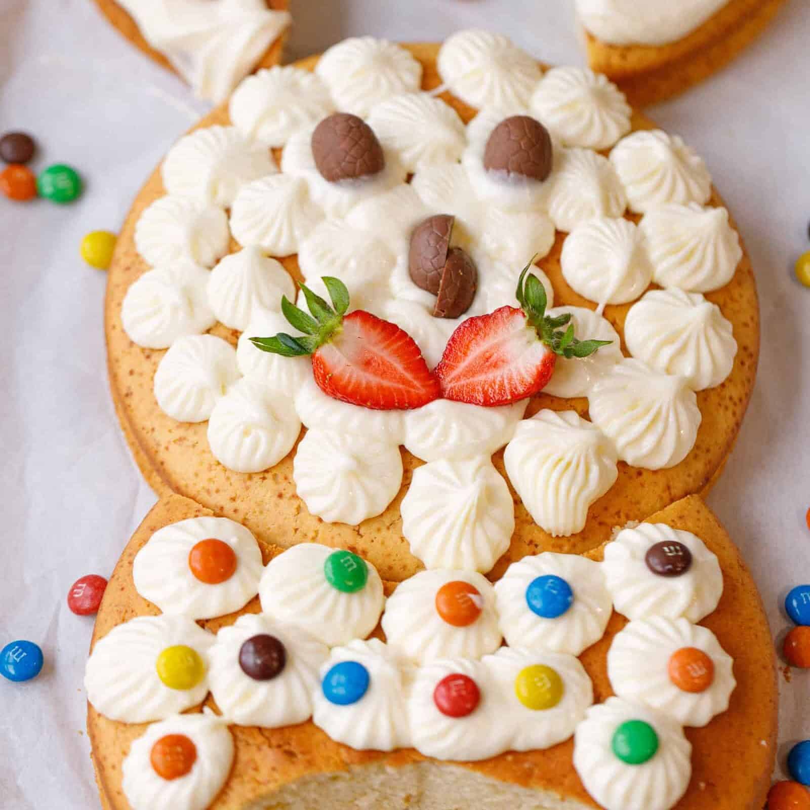 Close-up of a bunny-shaped Easter cake decorated with piped frosting, chocolate candy eyes, and strawberry slices for a nose.