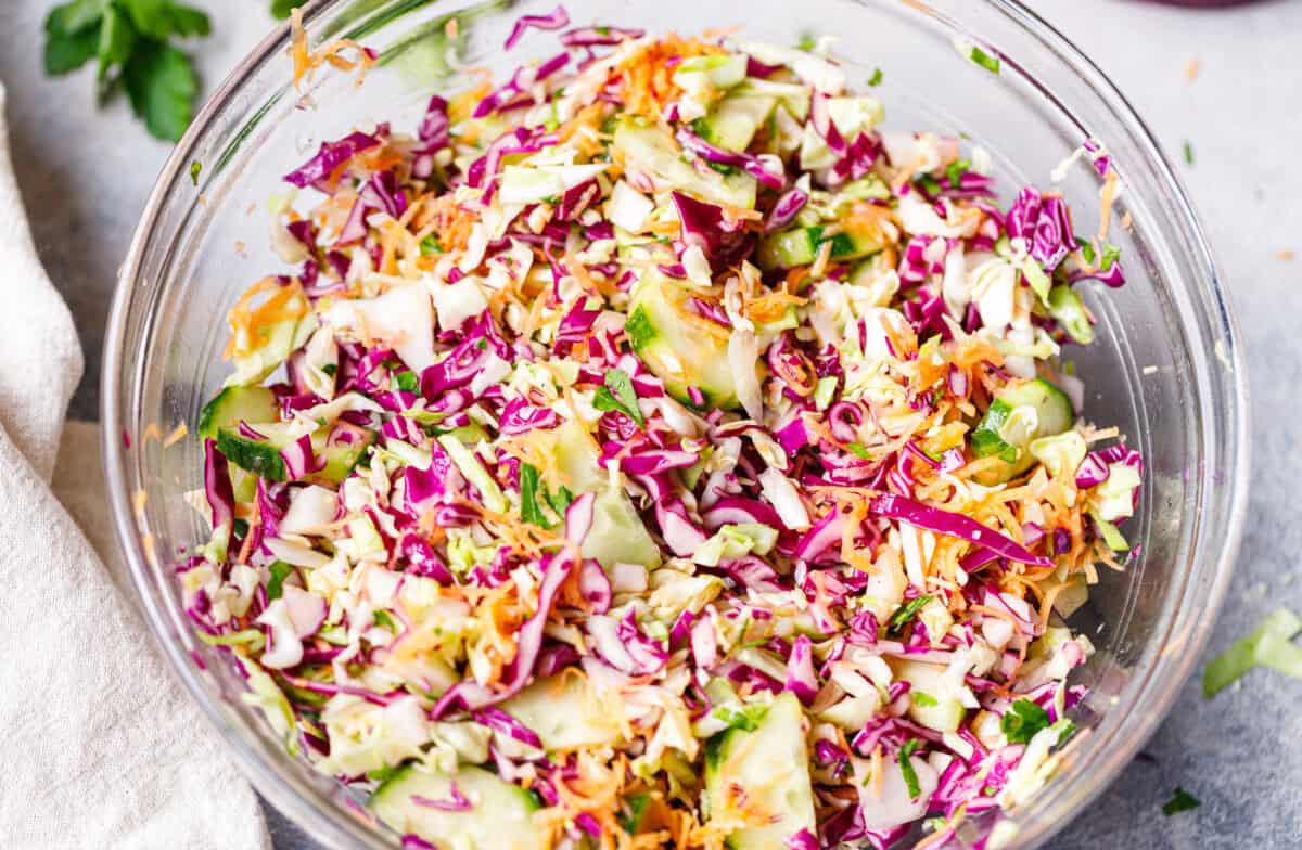Fresh cabbage salad in a glass bowl with green and purple cabbage, cucumber, carrots, and herbs