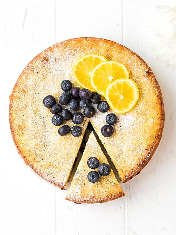Keto lemon cake with blueberries and lemon slices on top, dusted with powdered sweetener, one slice cut out, on a white surface