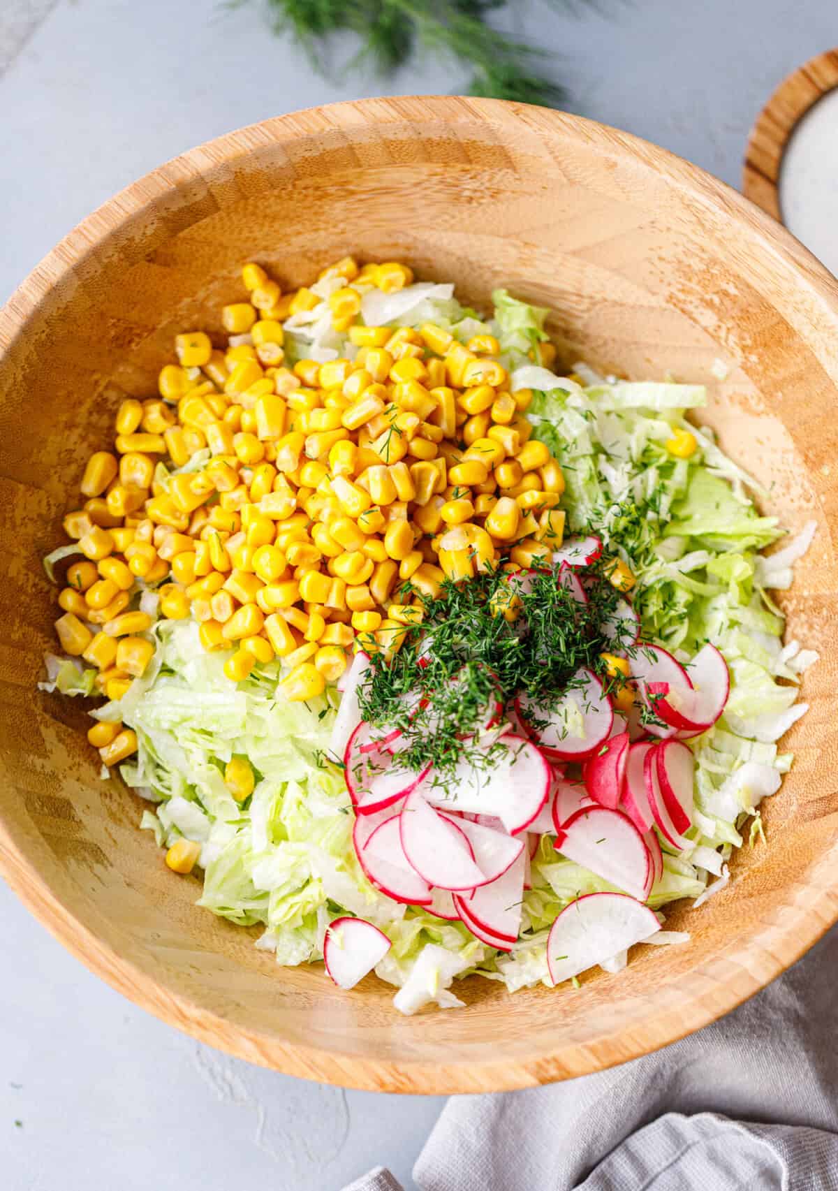 Overhead view of iceberg lettuce salad with corn, radishes, and fresh dill in a wooden bowl