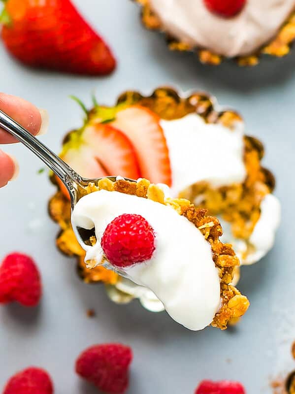 A spoon scooping a bite from a granola yogurt tart topped with a fresh raspberry, with a second tart showing a chocolate yogurt filling in the background.