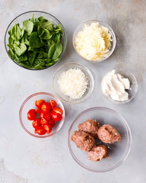 Ingredients for ground turkey pockets in glass bowls: fresh spinach, shredded mozzarella, parmesan, cream cheese, cherry tomatoes, and seasoned ground turkey