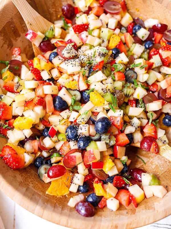 Close-up of fresh fruit salad with chia seeds and honey lime dressing in a wooden bowl — strawberries, blueberries, grapes, kiwi, orange, and diced apple tossed with fresh mint