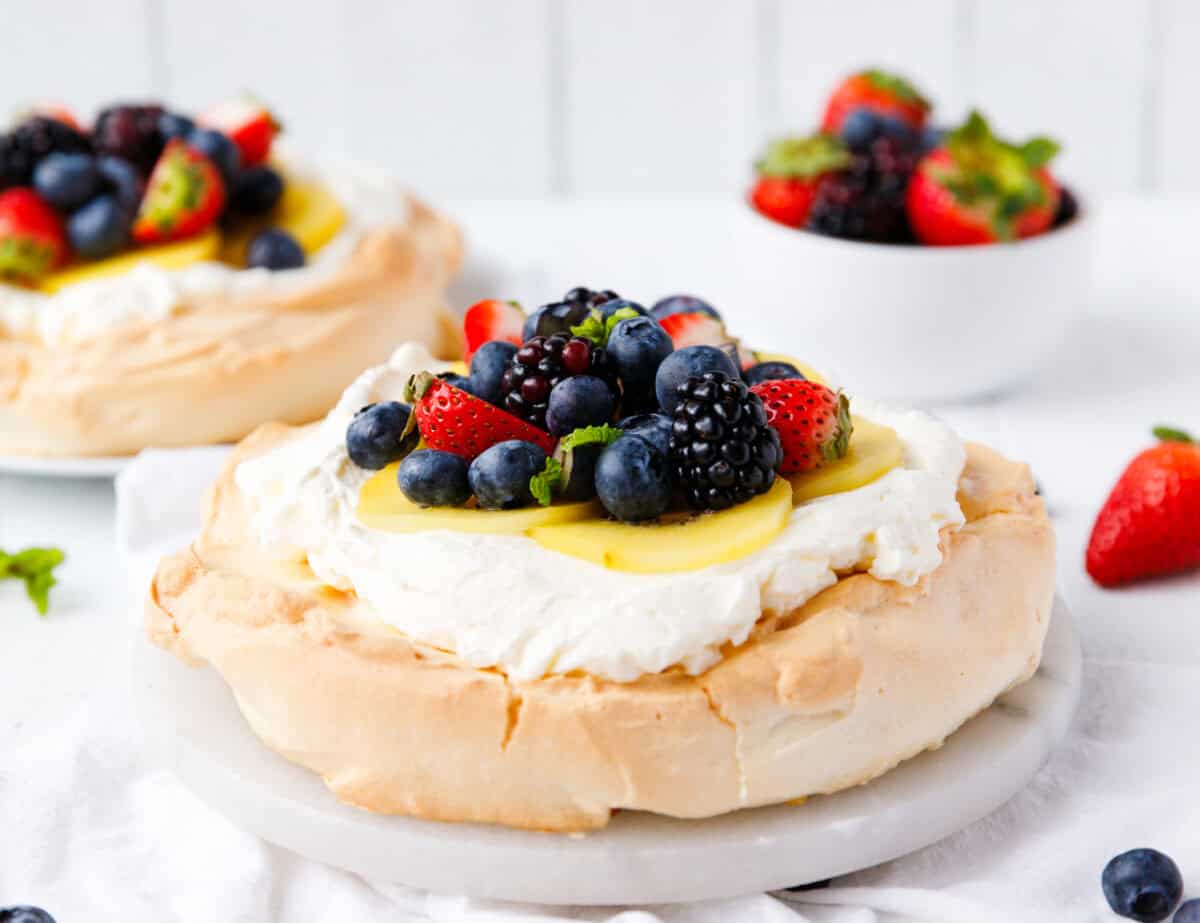 Two keto mini pavlovas on marble boards topped with whipped cream, kiwi slices, blueberries, blackberries, and strawberries, with a bowl of berries in the background
