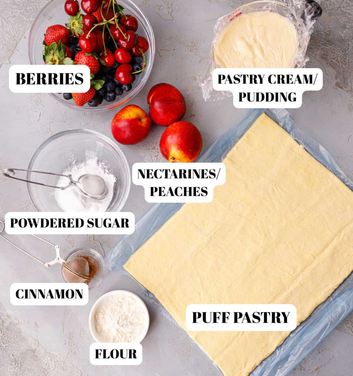 Labeled ingredients for puff pastry tart — puff pastry sheet, pastry cream or pudding, fresh berries, nectarines or peaches, powdered sugar, cinnamon, and flour on a gray surface