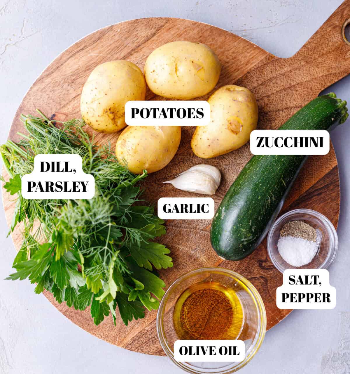 Labeled ingredients for zucchini and potatoes on a wooden board: potatoes, zucchini, garlic, dill, parsley, olive oil, salt, and pepper
