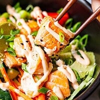 Close-up of chopsticks holding a piece of cooked salmon with spicy mayo and sesame seeds above a poke bowl