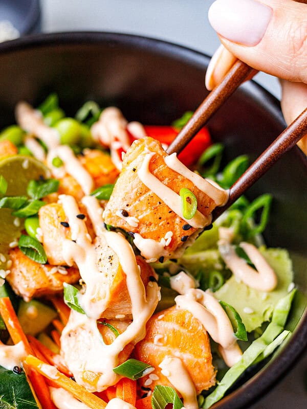 Close-up of chopsticks holding a piece of cooked salmon with spicy mayo and sesame seeds above a poke bowl
