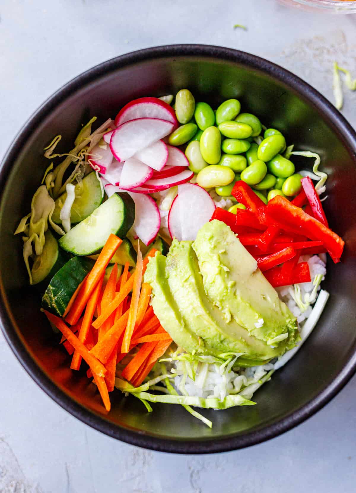 Poke bowl base of rice topped with cabbage, cucumber, carrot, radish, edamame, red pepper, and sliced avocado before adding salmon