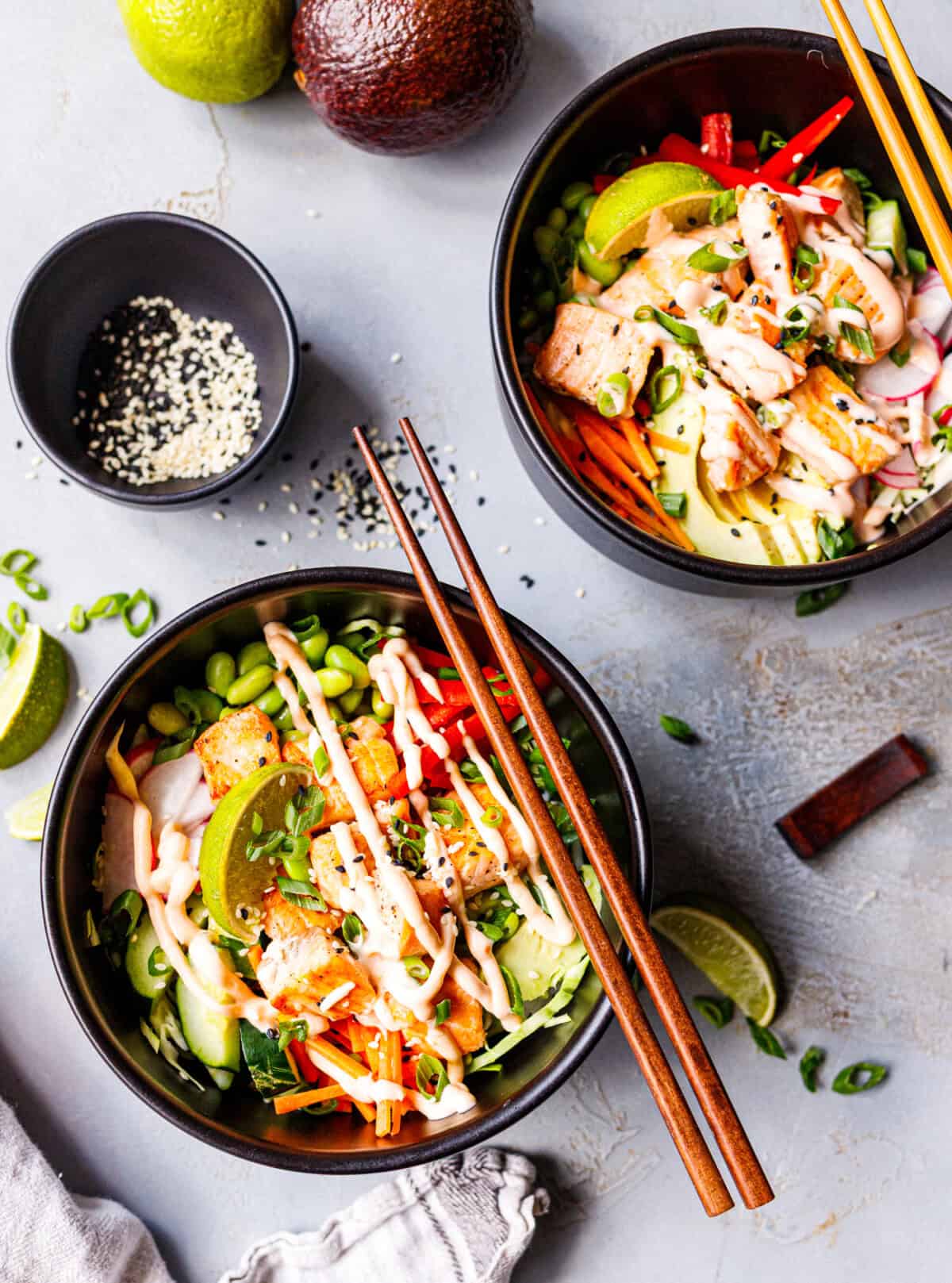 Two cooked salmon poke bowls seen from above with seared salmon, spicy mayo, edamame, red pepper, carrot, cucumber, avocado, and lime, wooden chopsticks resting on the rim