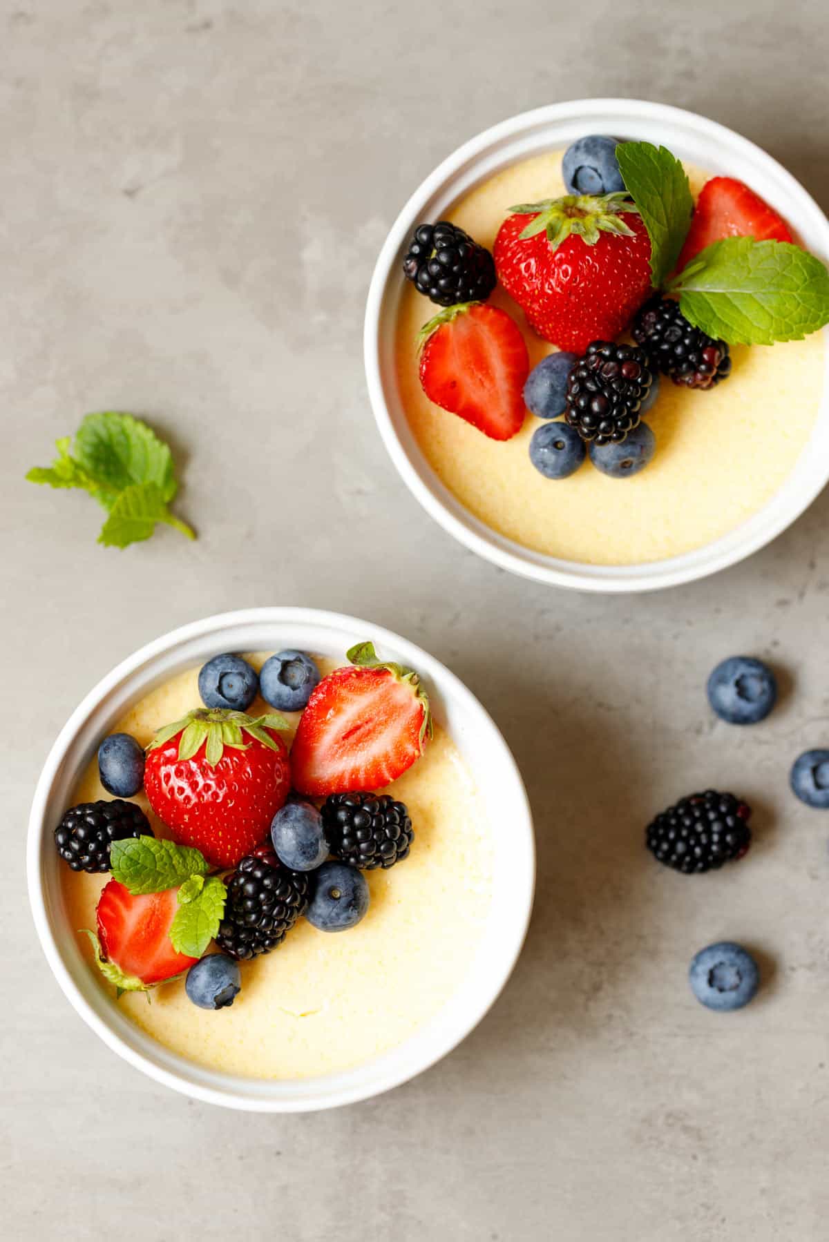 Overhead view of two keto custard cups garnished with strawberries, blueberries, blackberries and mint on a gray background