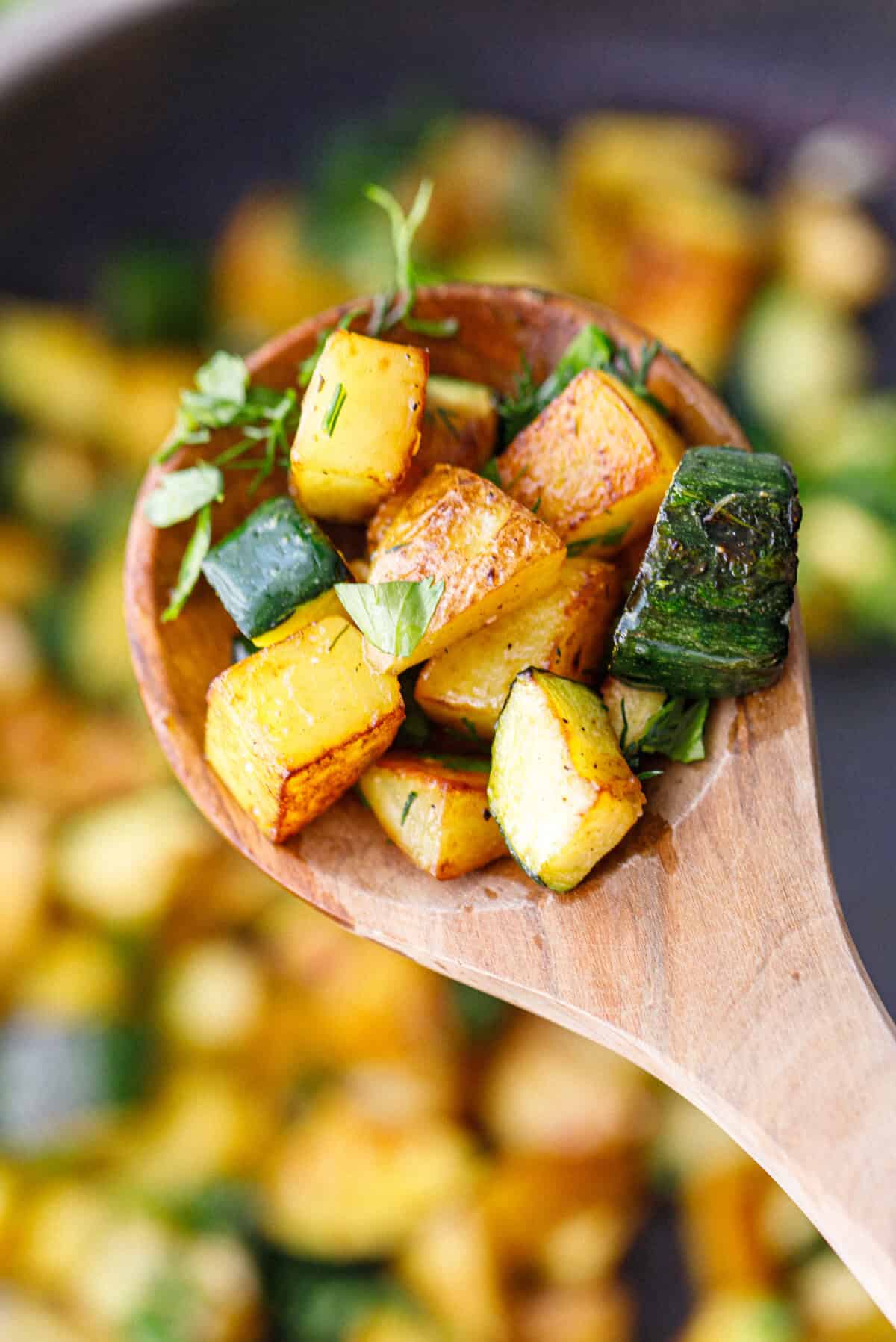 Close-up of a wooden spoon lifting golden pan-cooked zucchini and potatoes topped with fresh dill and parsley