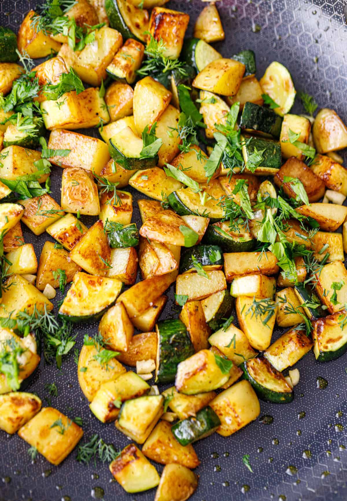 Overhead view of a skillet full of browned zucchini and potatoes garnished with dill and parsley