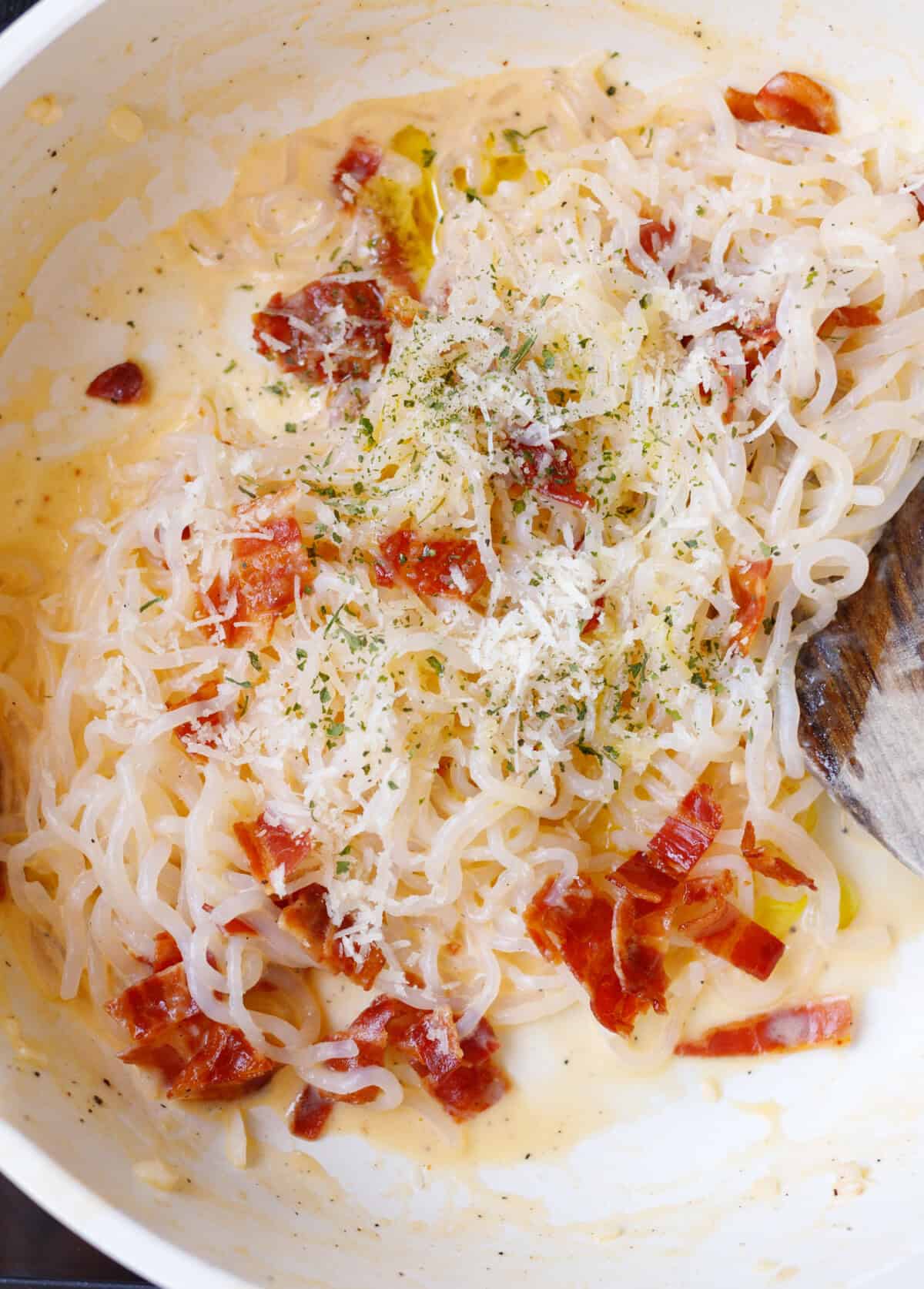 Konjac carbonara in a pan topped with grated parmesan, bacon, and parsley before serving