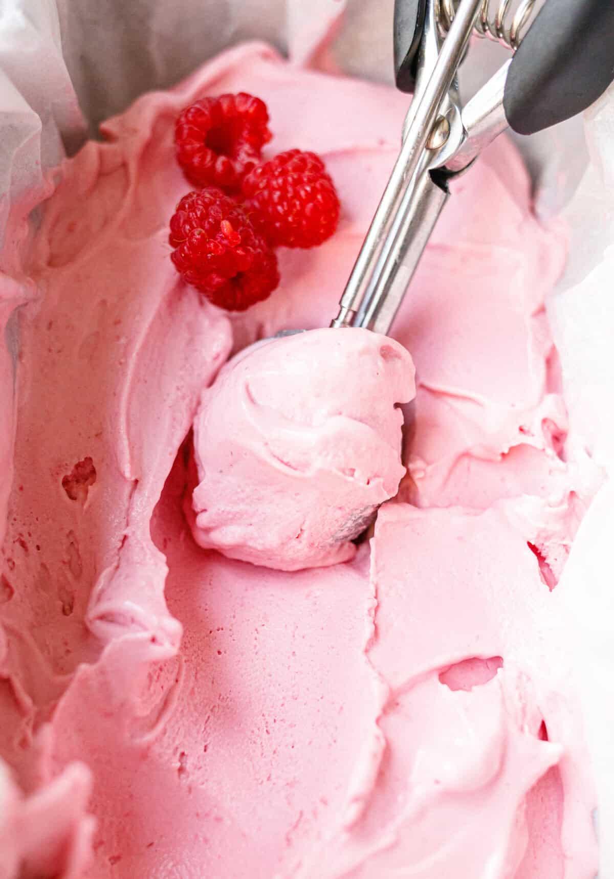 Scooping low-carb raspberry mascarpone ice cream from a parchment-lined container with fresh raspberries