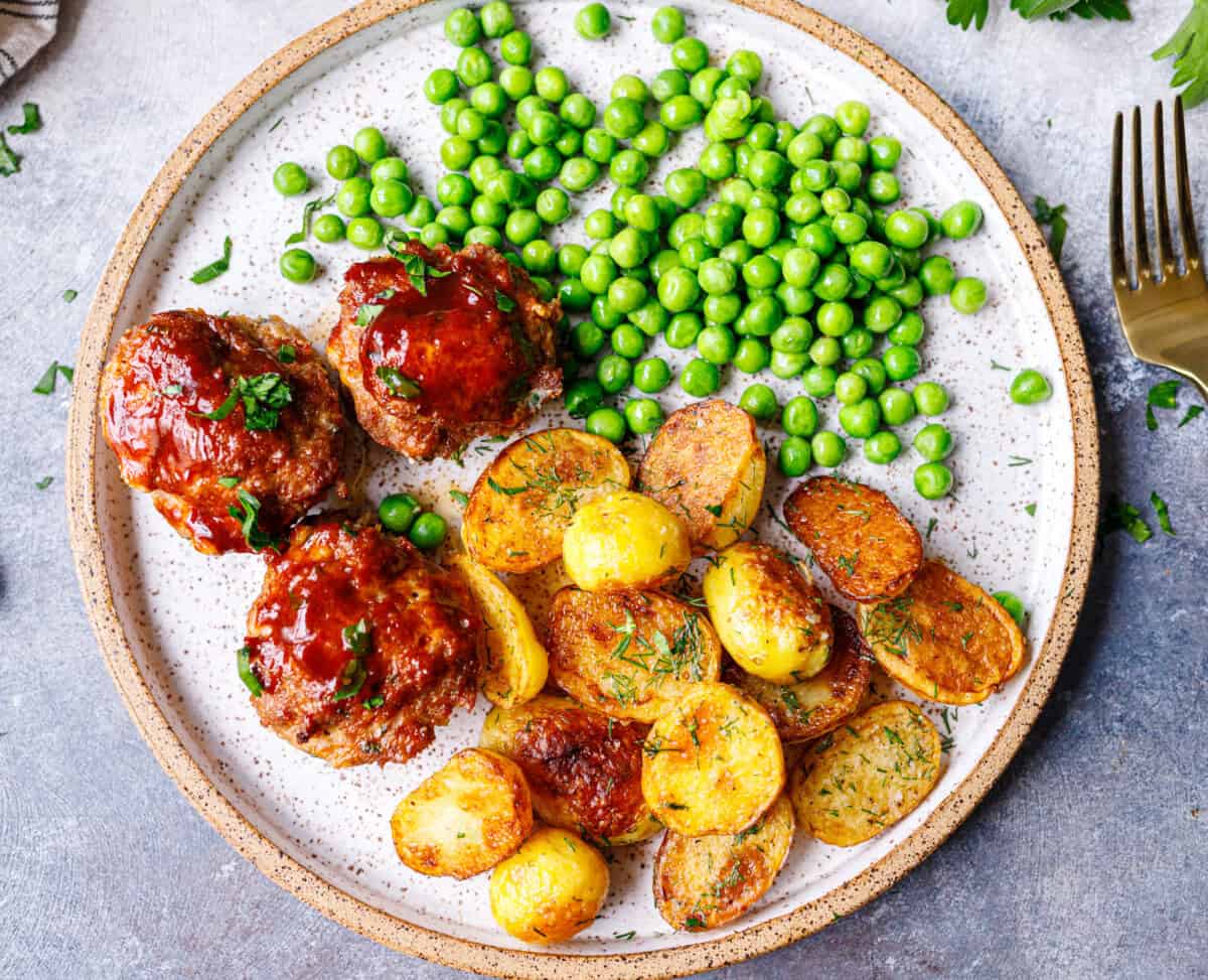 Three mini meatloaf muffins with BBQ glaze on a speckled plate with dill potatoes and green peas