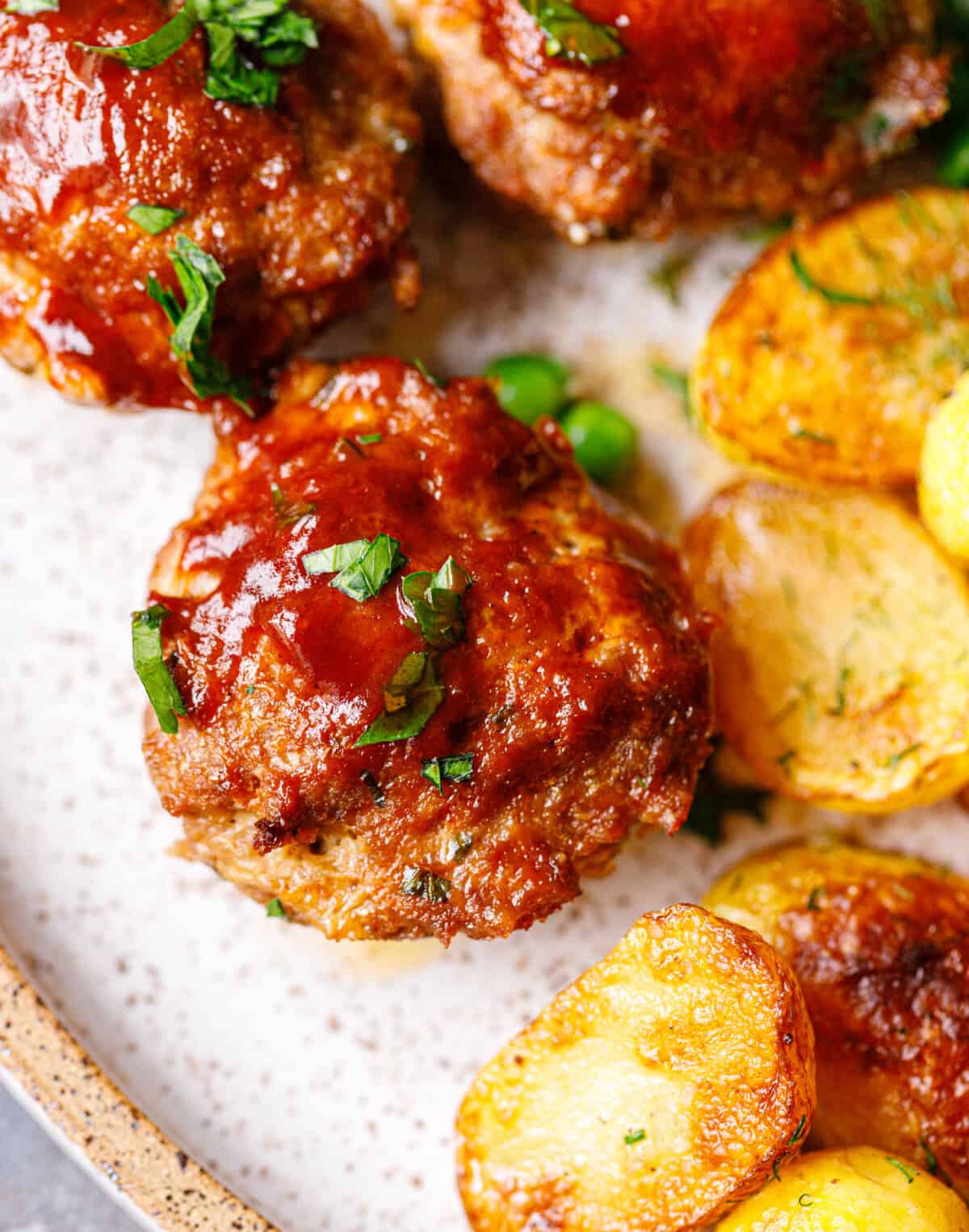Close-up of a BBQ-glazed mini meatloaf topped with parsley, next to crispy dill potatoes and green peas