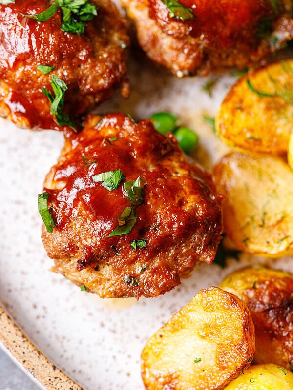 Close-up of a BBQ-glazed mini meatloaf topped with parsley, next to crispy dill potatoes and green peas