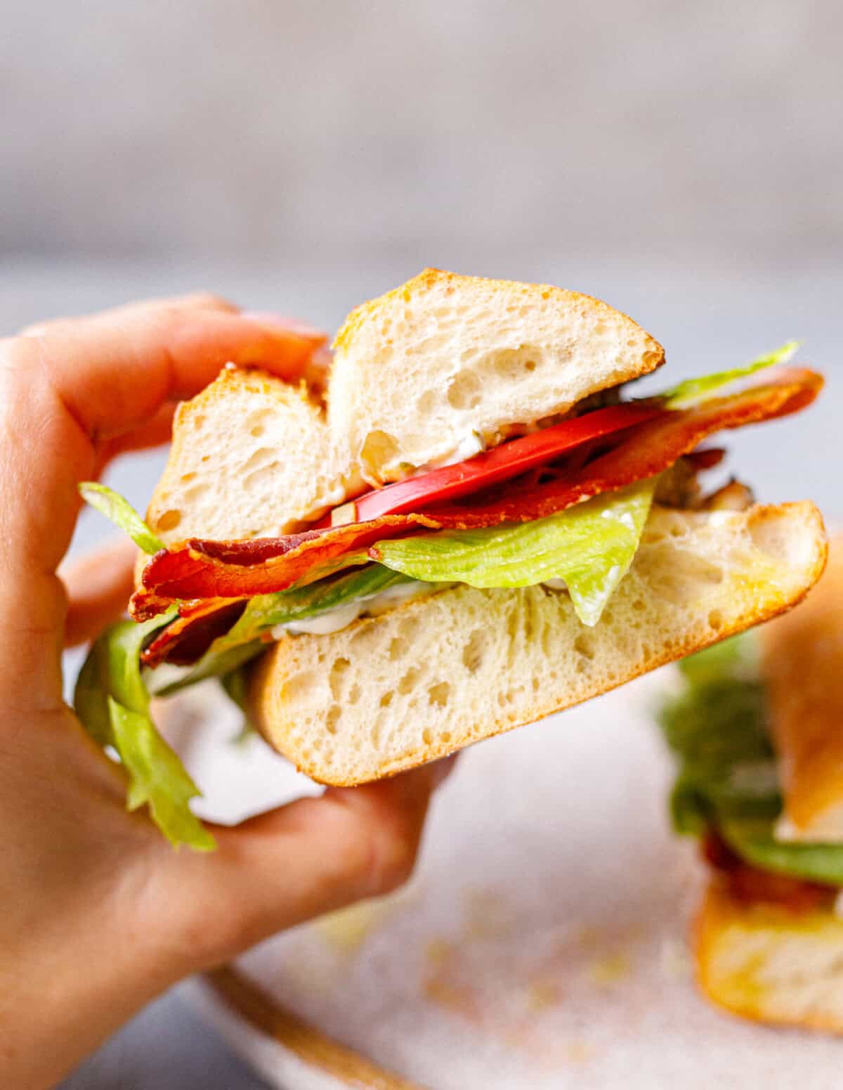 Hand holding a ciabatta BLT sandwich showing layers of crispy bacon, tomato, lettuce, and pickle mayo