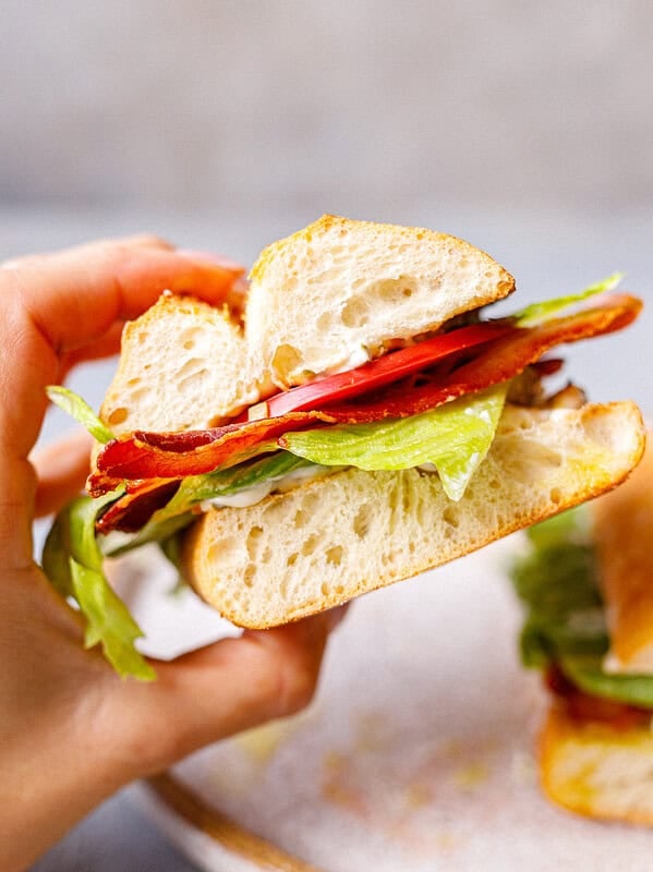 Hand holding a ciabatta BLT sandwich showing layers of crispy bacon, tomato, lettuce, and pickle mayo