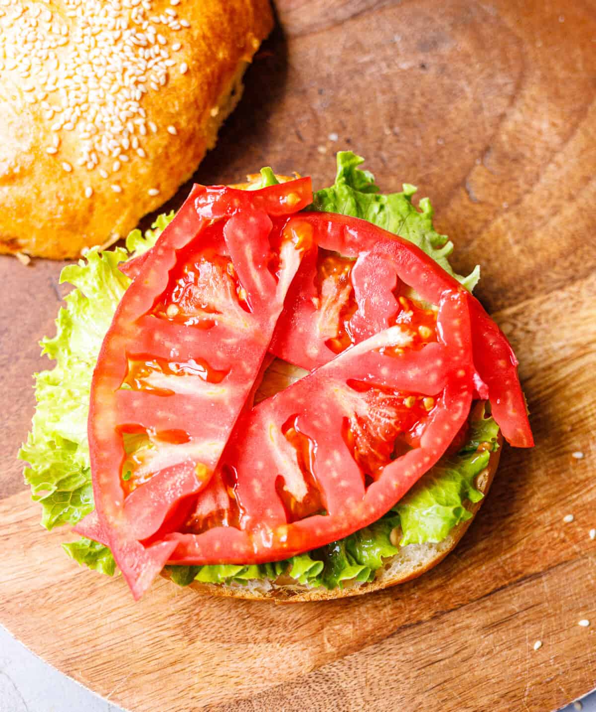 Sliced tomato layered over lettuce on the bottom half of a sesame bun